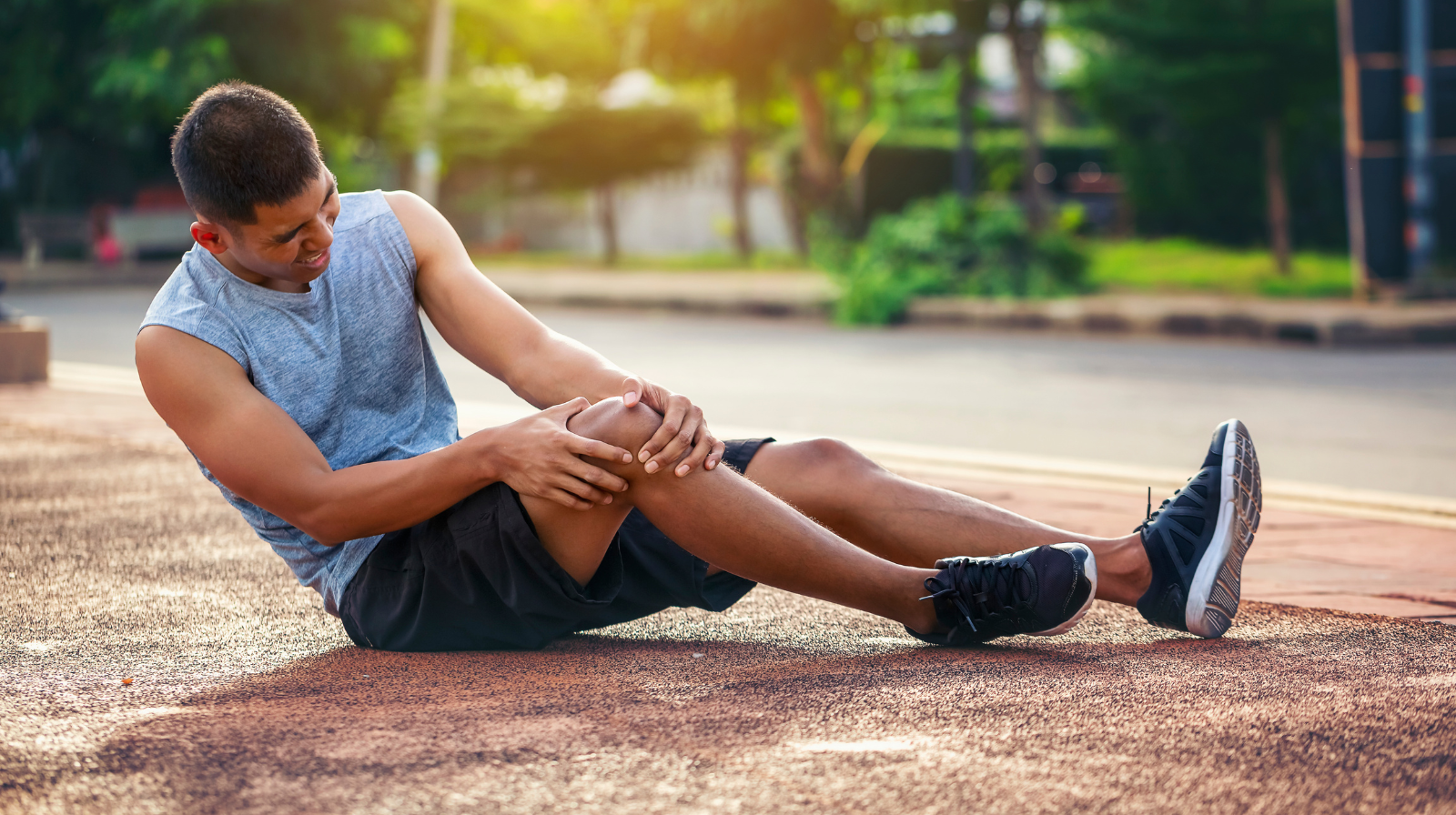 Homem sentado no chão de uma pista de corrida segurando o joelho após uma torção